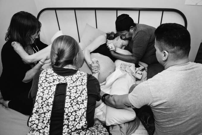 A mother in pain in active labor on her bed during a home birth surrounded by her husband and birth team.